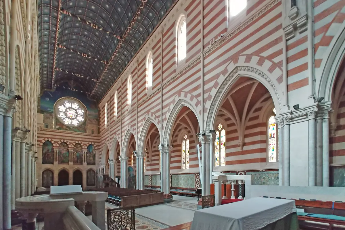 Boční pohled do interiéru kostela z galerie, s pruhovanými stěnami, modrým stropem se vzorem hvězd, lomenými oblouky a obkládanou podlahou v červené a zelené barvě.