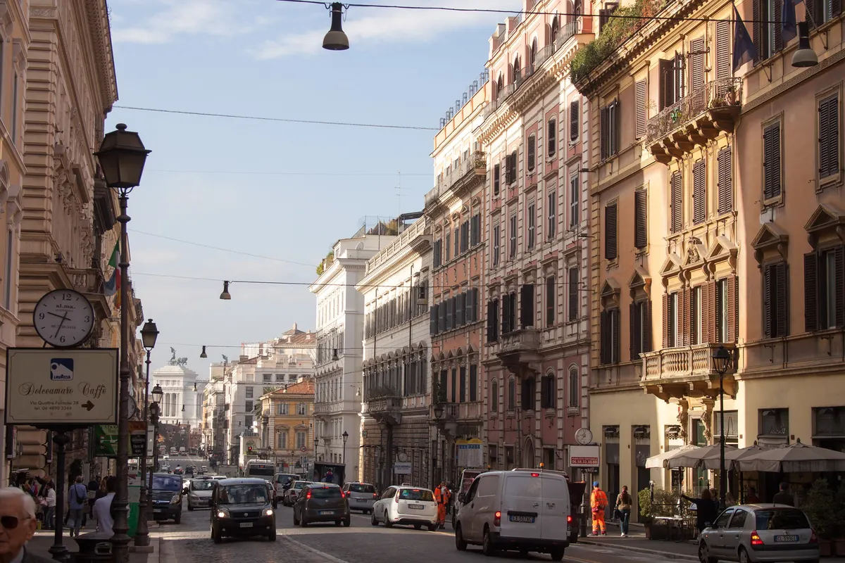 Via Nazionale v Římě v odpoledním světle, dlouhá ulice s vysokými okrovými a růžovými paláci s balkony, směřující k bílému monumentu v dálce.